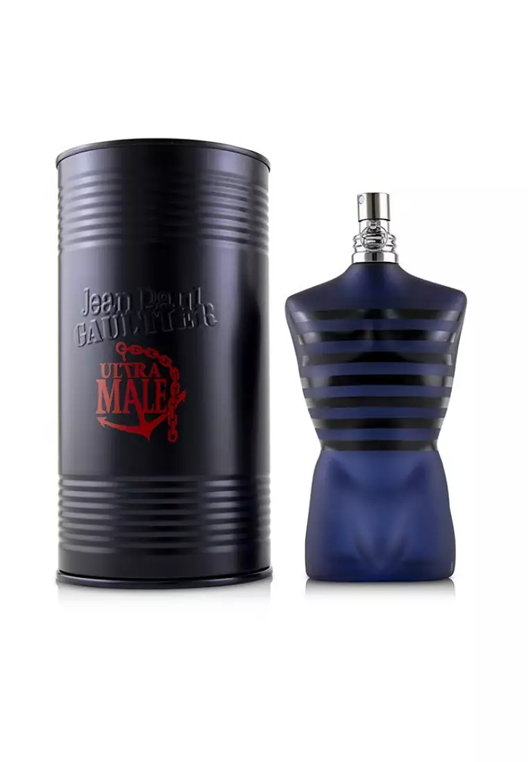 Jean Paul Gaultier Ultra Male fragrance shown with a black ribbed cylindrical tin and a dark blue bottle shaped like a male torso with horizontal black stripes, on a white background.